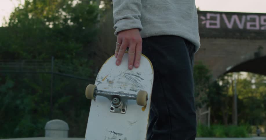 A skateboarder taps his board on the ground while standing near an overpass