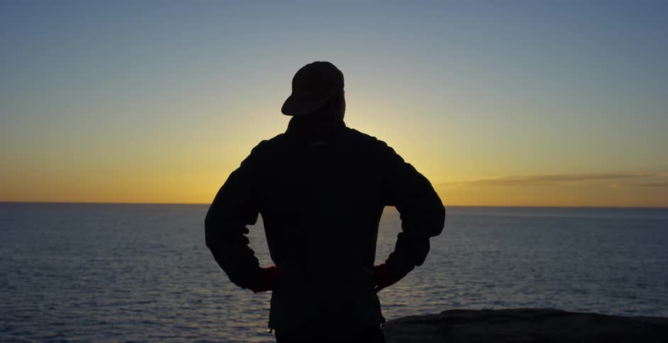 A man is standing by the ocean watching the sunset