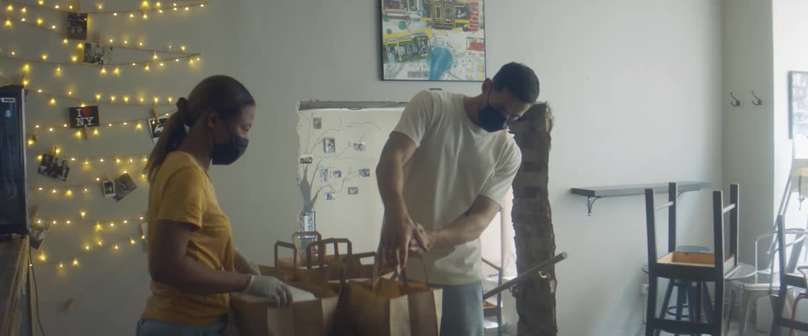 A man and a woman with mask in a room with brown paper bags with a wall in the background