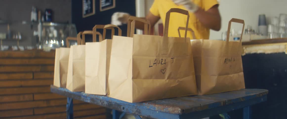 Woman fills labeled paper bags with food orders indicating correct order to man who arrive to pick an order up