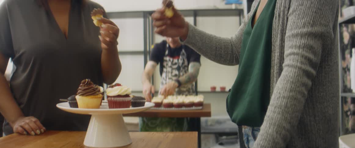 Two women sample desserts while man arranges cupcakes on table in background