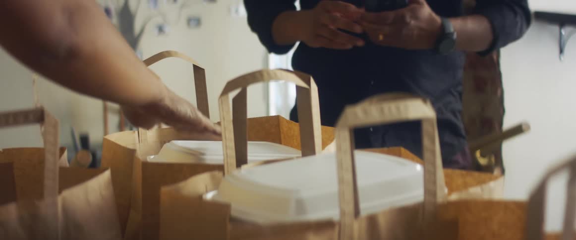 A person in a room putting a styrofoam container into a brown paper bag as a man with a mask takes a brown paper bag