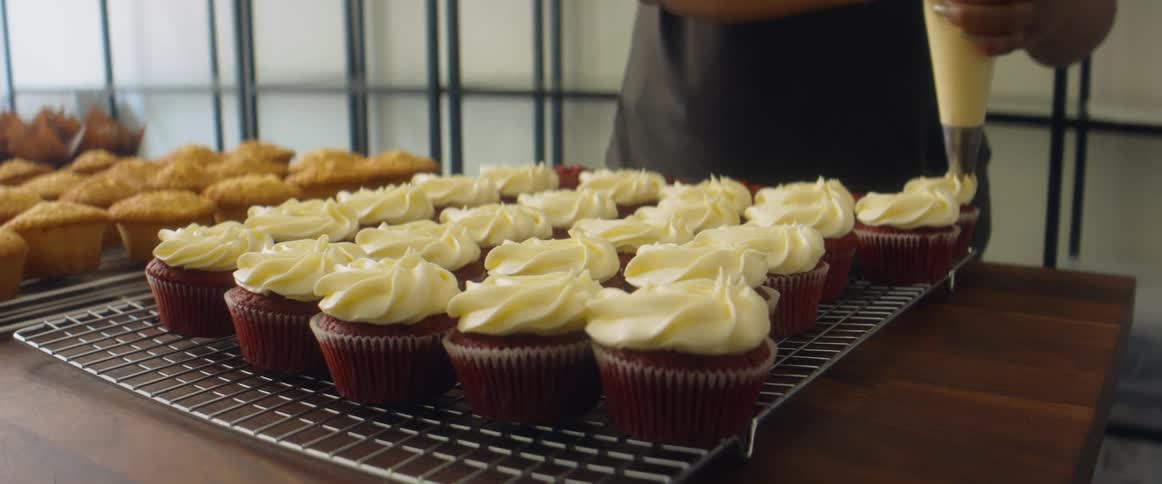 A person in a bakery putting frosting on top of cupcakes on a table
