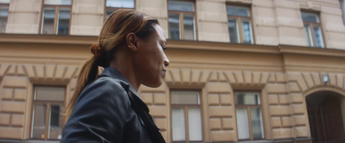 A woman with a bag and jacket walking a city street with buildings the sky and clouds in the background