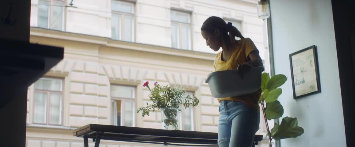 A woman in a room wiping a table and adjusting a flower in a vase next to a window