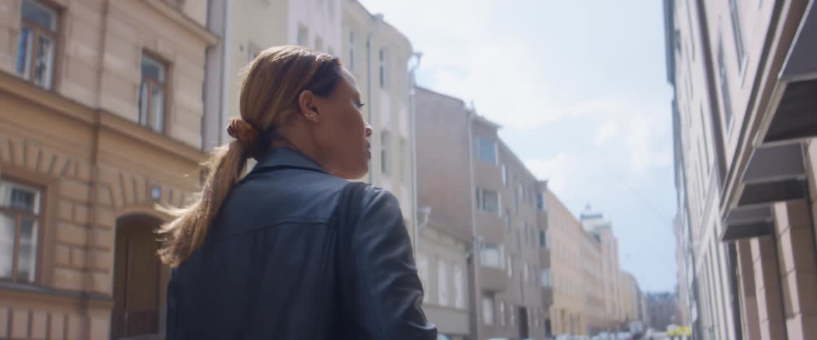 A woman walking in a city street with vehicles surrounded by buildings with the sky and clouds in the background