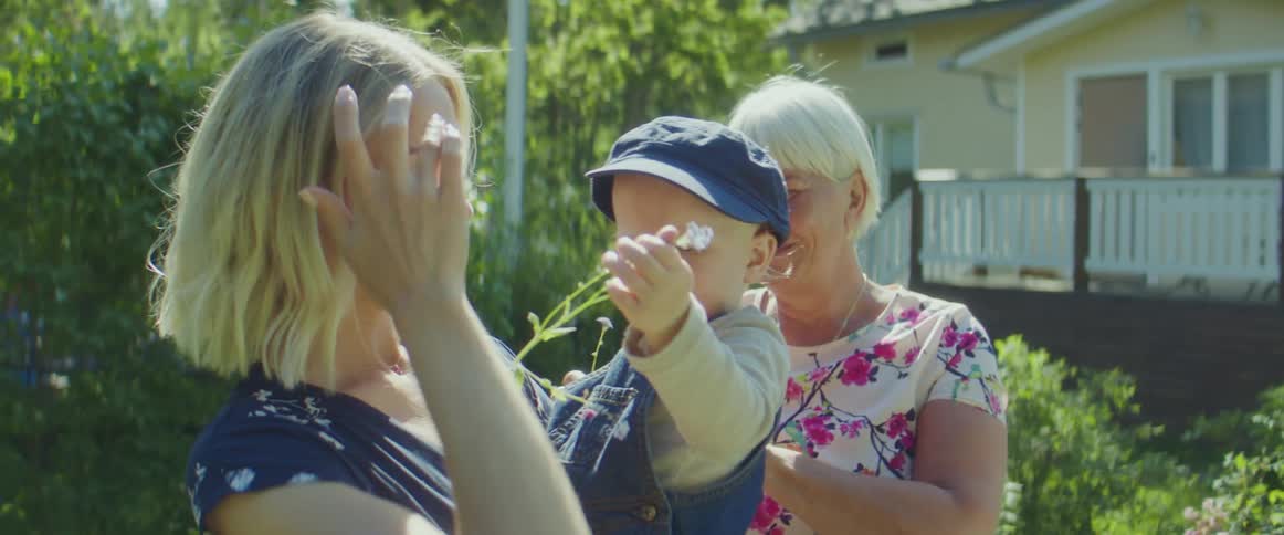 Mom smiles as baby grabs the flower from her hair as grandma looks as they all stand in the garden