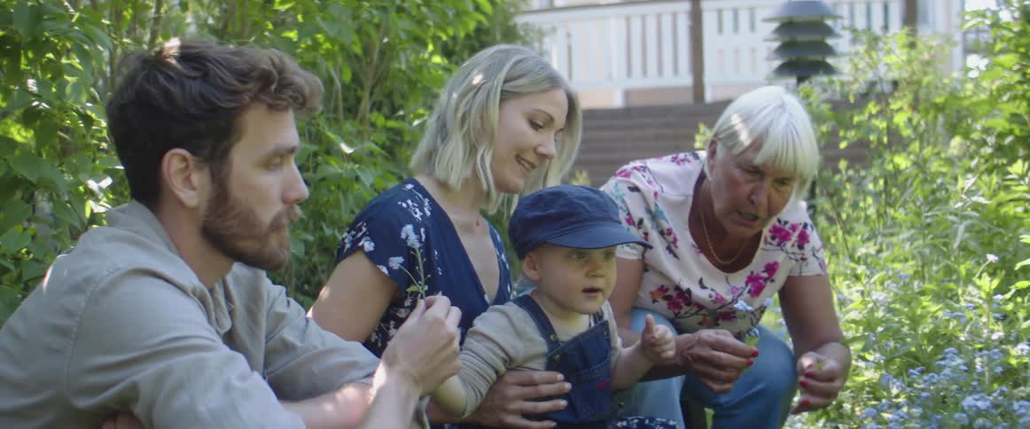 A woman holds her toddler as an elderly woman picks flowers in the garden and a man shows them to the toddler