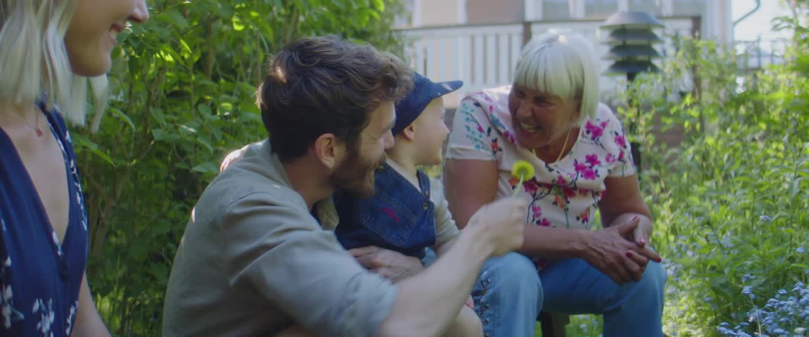 A man holding a small boy and a flower leans over to let the woman smell it