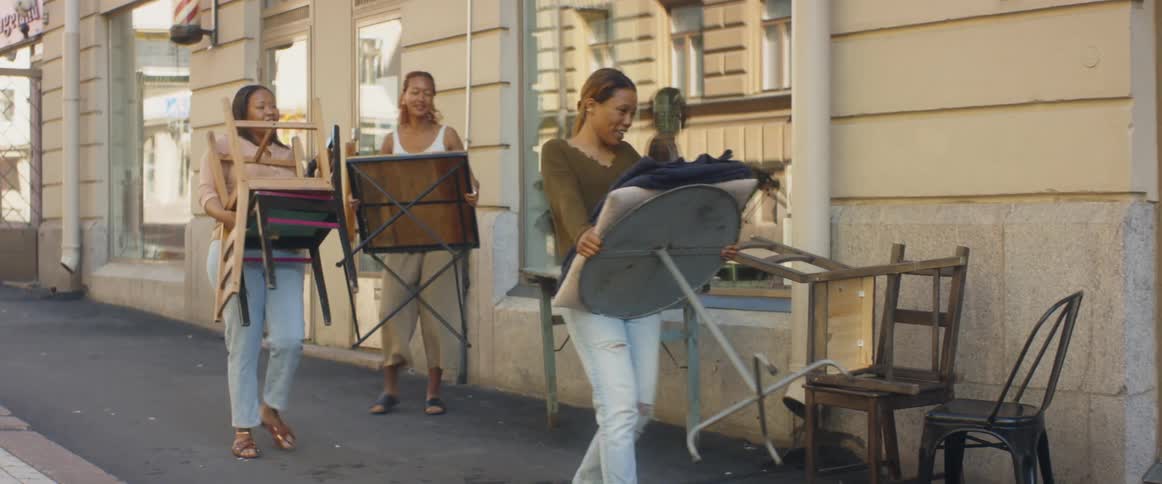 Three women next to a building in a city street moving chairs and tables