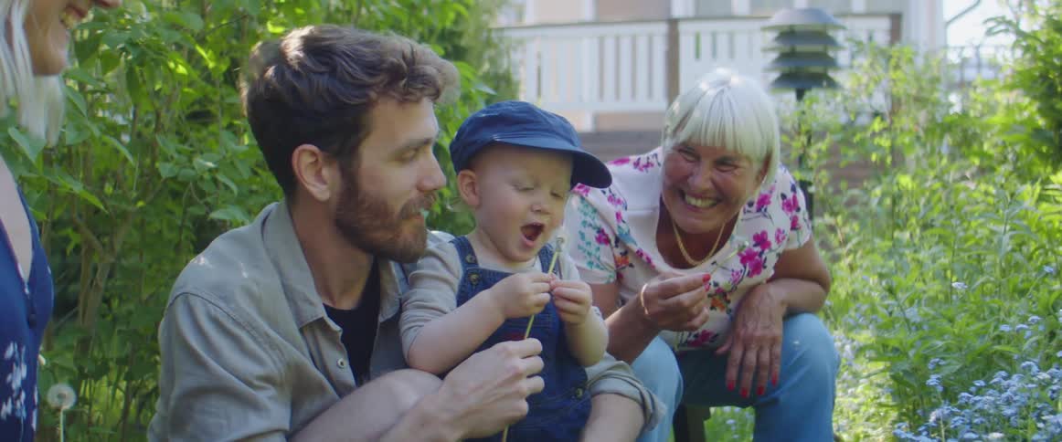 A man a boy and two women with the man blowing on dandelions with a house in the background