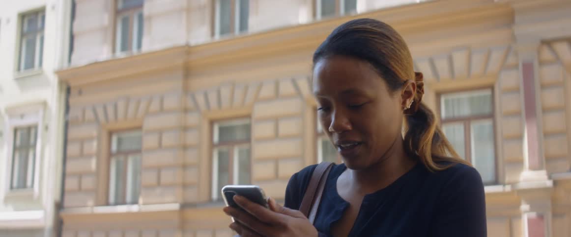 A woman with a bag walking on a city street and pulling out a smart phone with a building in the background