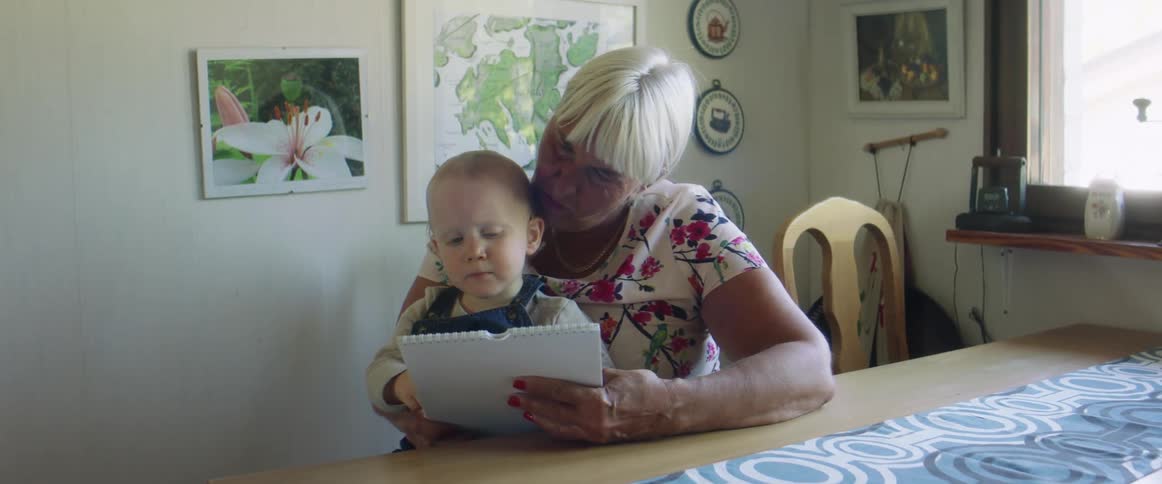 A grandmother reads with her grandson at a kitchen table on a sunny day