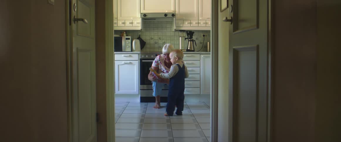 An elderly woman kneels and shows a toddler a story book who then approaches her to look at the book