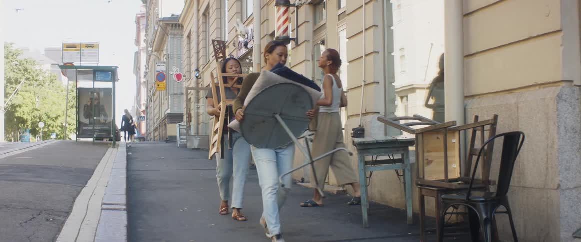 Three women hold outdoor furniture and place it in front of a storefront
