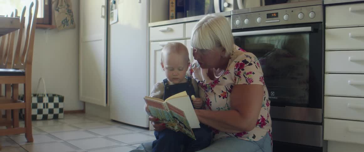 Grandmother sits on kitchen floor near stove with grandson on her lap while she reads him a book