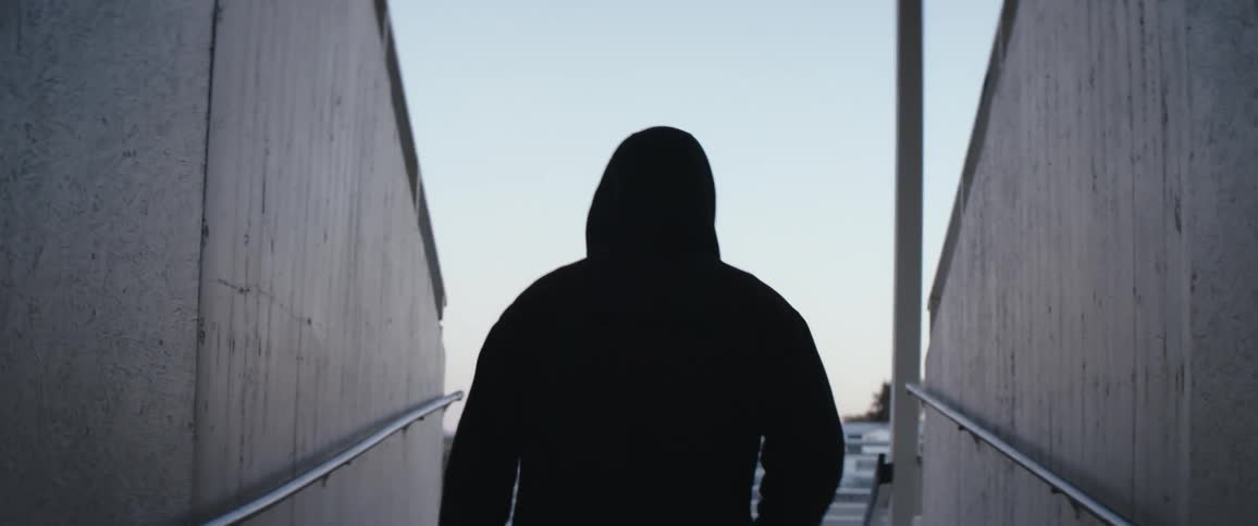 A person with a hoodie walking in a corridor with trees the sky and clouds in the background