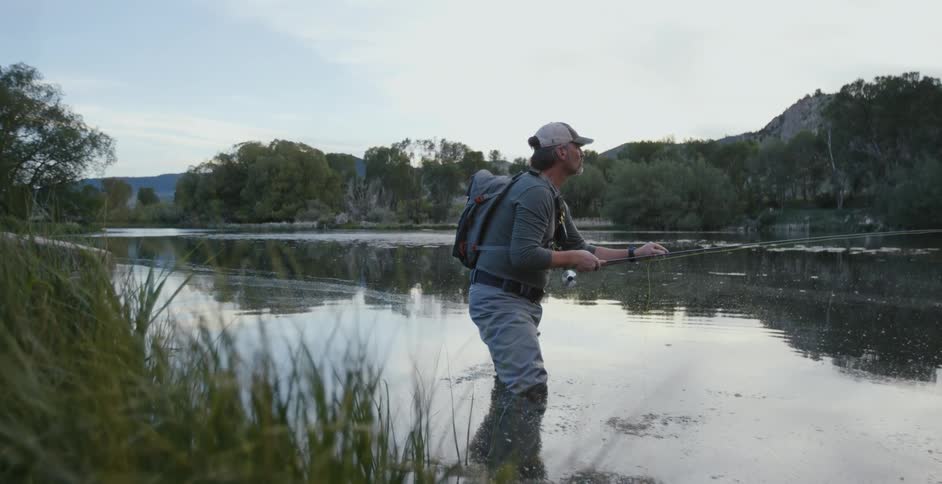 A man with a hat and a backpack fishing with a fishing rod in a river with the sky and clouds in the background