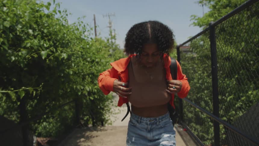 A teen girl walking by trees and a chain link fence with a backpack