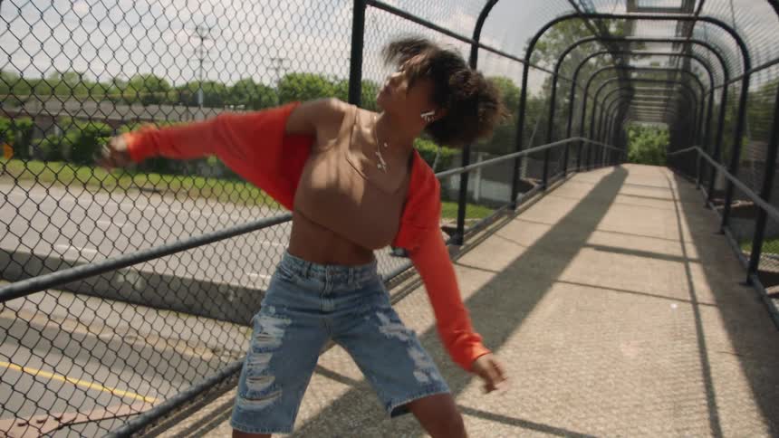 A young woman performs dance moves while she is on a walking bridge over a roadway
