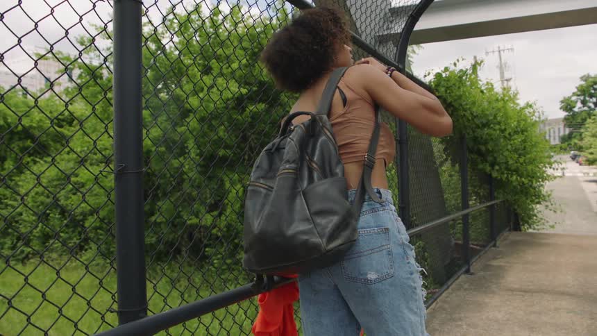 A woman on a bridge lifts her backpack from the ground and places it over her shoulder before grabbing her jacket from a handrail and continuing across the bridge
