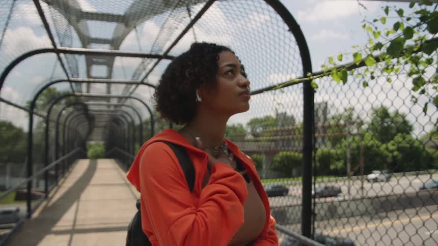 A young woman standing on a walking bridge over traffic holds her chest while breathing deliberately