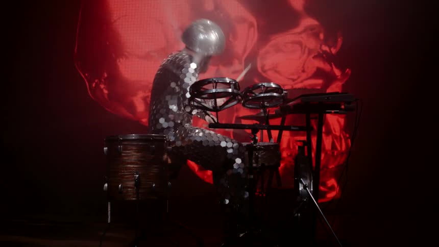 A musician on stage beats his drums and shakes his head in front of a revolving red skull