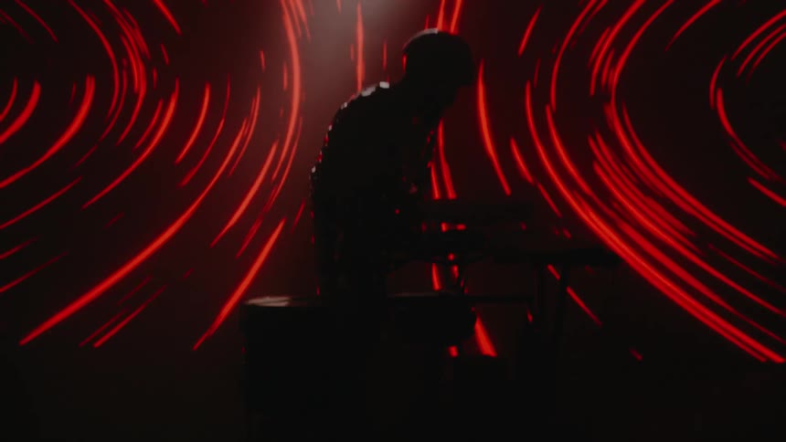 A man in a room playing drums with drumsticks with a screen in the background