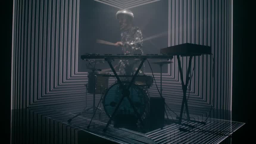 A man in a room playing drums with drumsticks with a screen in the background