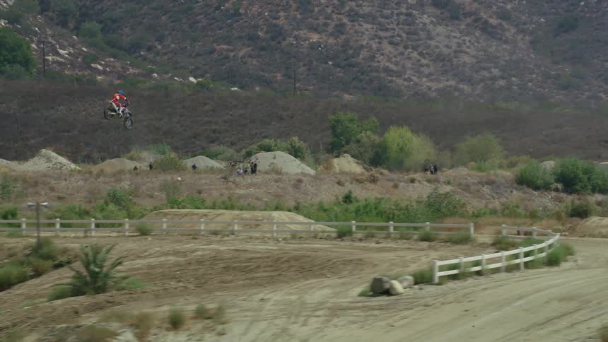 A person on a motorcycle rides up a dirt road in the countryside and flies through the air before landing on a ramp on the opposite side of a fence