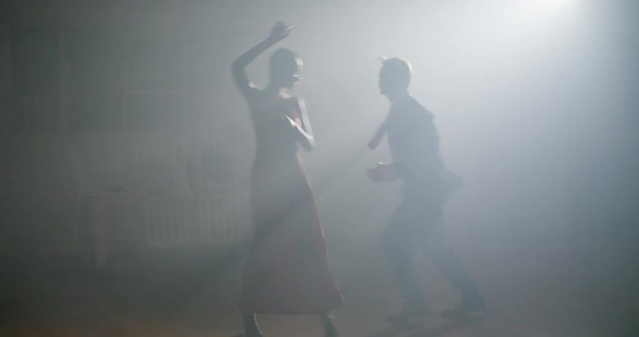 Man in suit and woman with short hair and red dress dance in smoke filled room