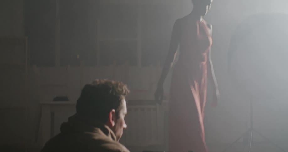A man on the floor with a camera filming a woman with a dress in a room with a wall in the background