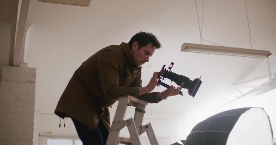 A photograph holds a camera while standing on a ladder in a studio