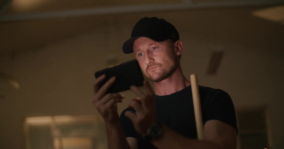 A man with a hat in a room looking at a smart phone with a wall in the background