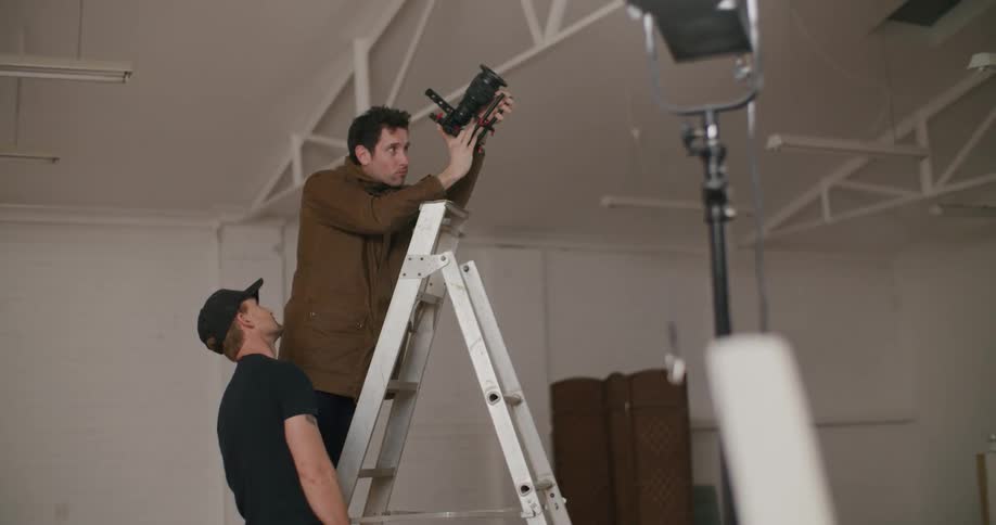 A photographer stands on a ladder and looks through his camera lens as another man stands behind him and watches