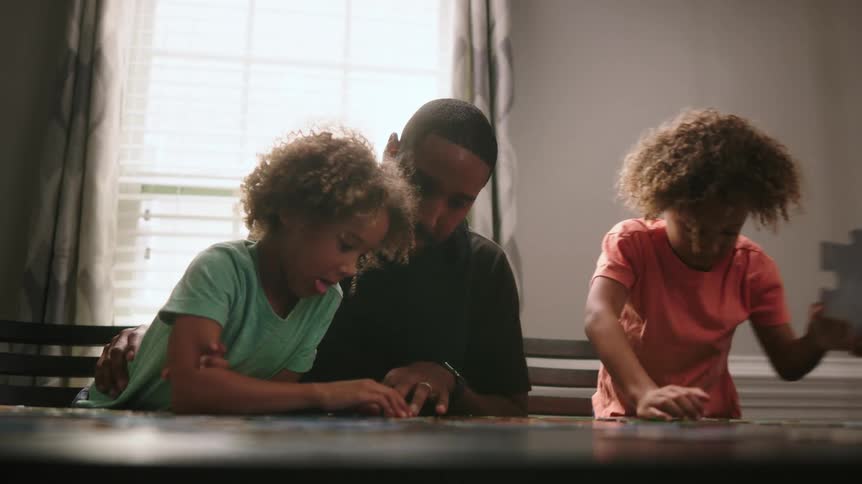 A father helps his two young daughters to assemble a large jigsaw puzzle on their table