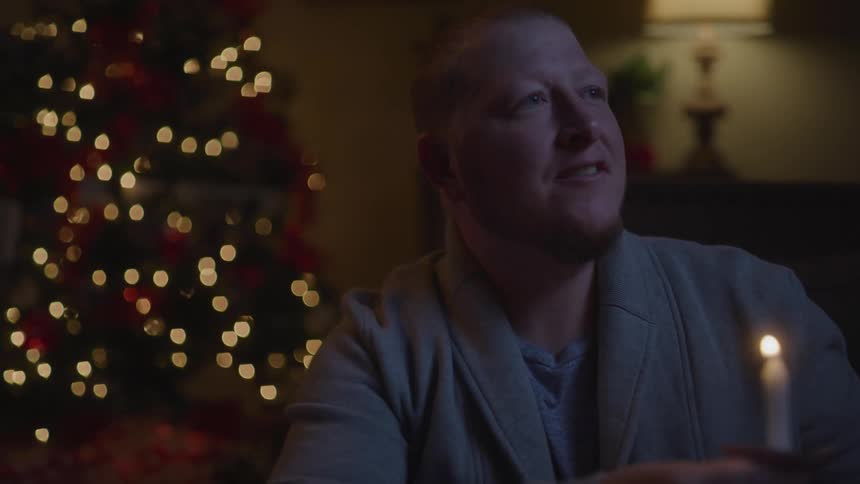 A man sits in front of a tree with lights while he lights and holds a small candle