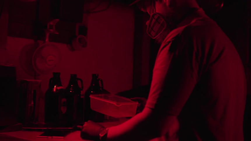 A man in a dark room wearing gloves and a mask moves around some liquid in a clear bin