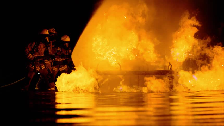 Three firefighters hold a hose and spray a fire burning bright orange at night
