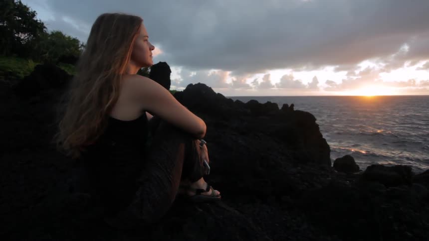 A blonde woman sits on a rock at sunset