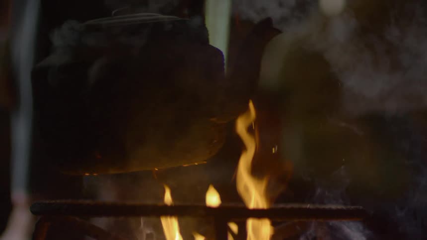 A man uses tongs to take a kettle off an open flame outside