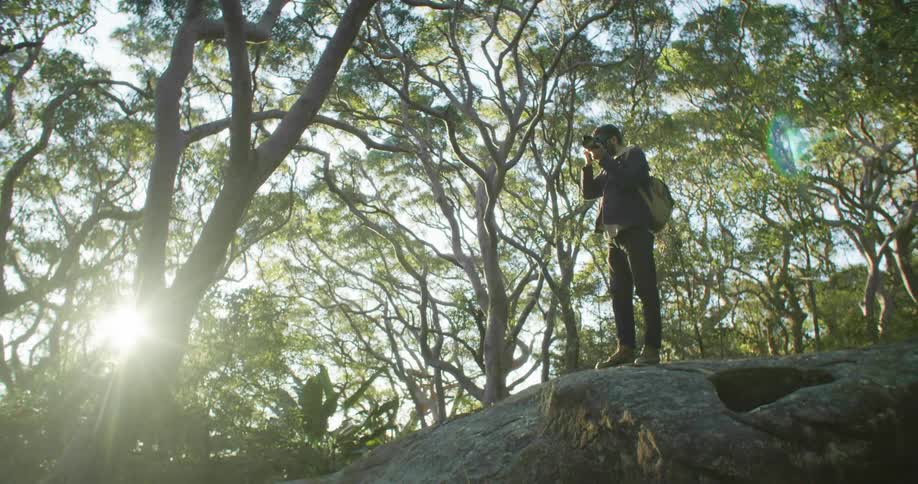 A man with a camera and a backpack in a forest taking a picture with the sky and clouds in the background
