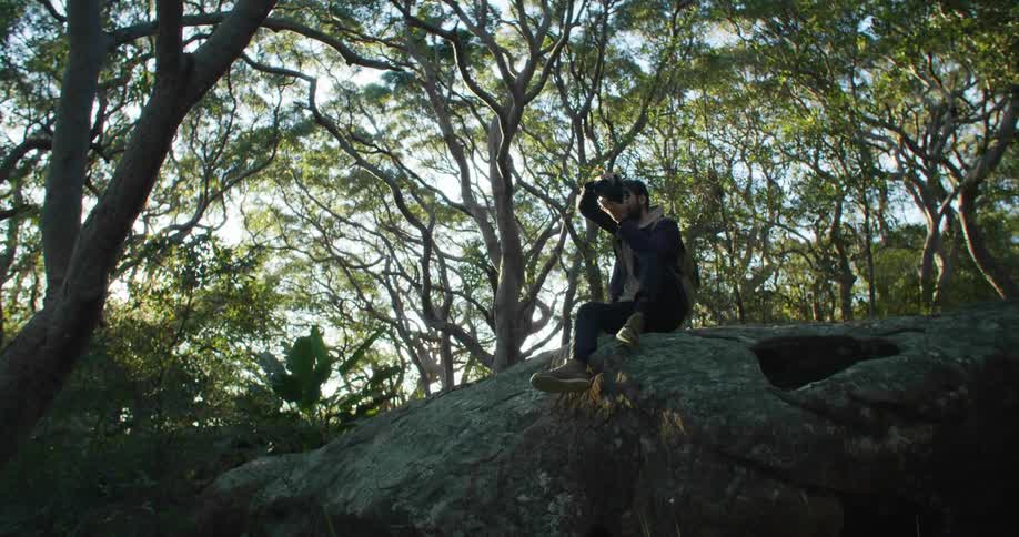 Man with a beard and a coat sits on a big rock and takes photos in the woods as the sun shines in the background