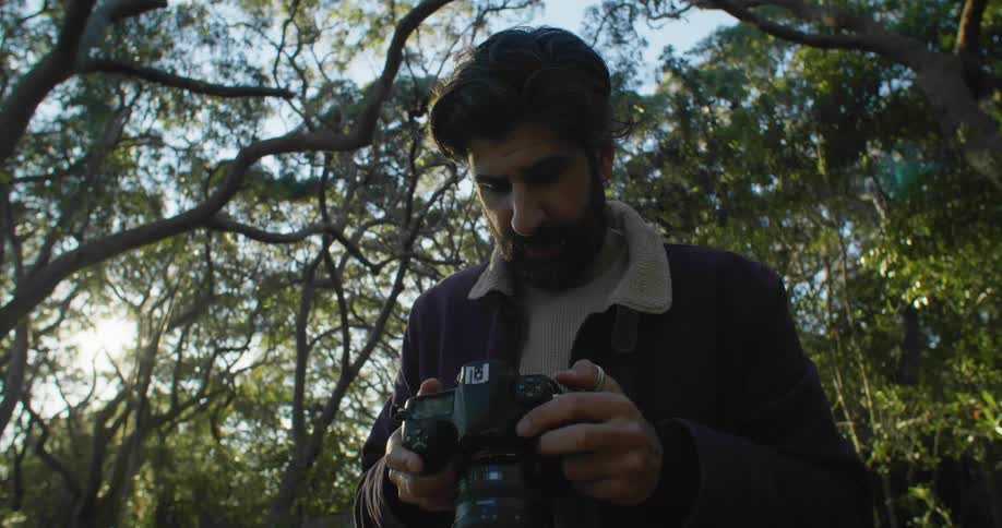 A man stands outside in a wooded area looking at a camera screen in his hand