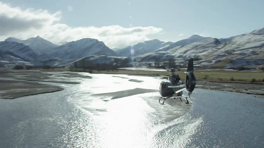 A helicopter flies over a shallow stream near snowtopped mountains