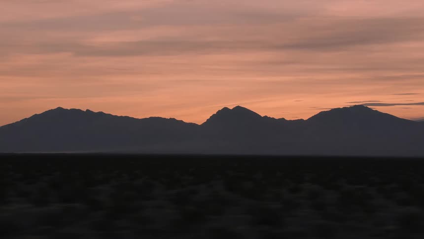 Distant mountains loom over a vast shrubland under a cloudy sky at sunset