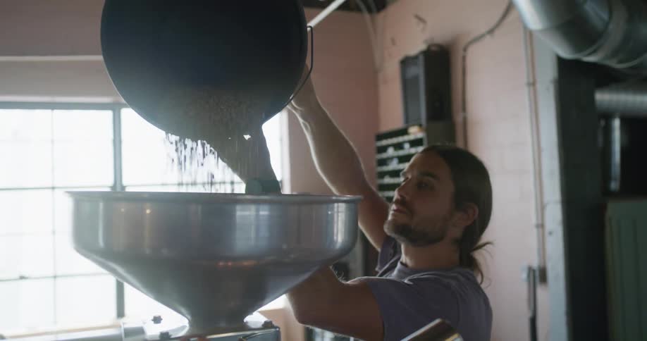 A man in a room pouring coffee beans from a bucket into a machine with a wall and window in the background