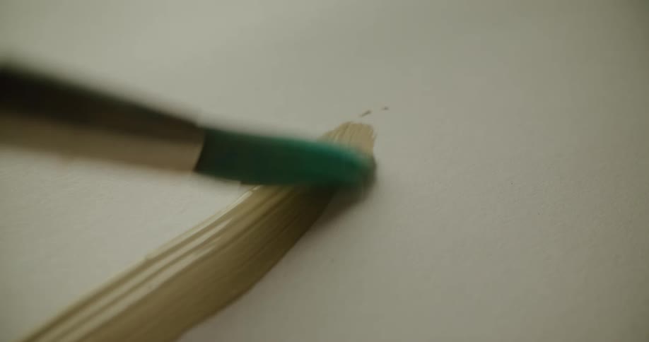 A green paint brush applies beige paint onto a white canvas