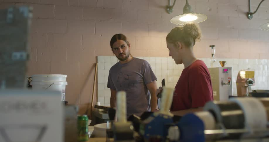 Man and woman talk while in a room with various machines and buckets and sacks