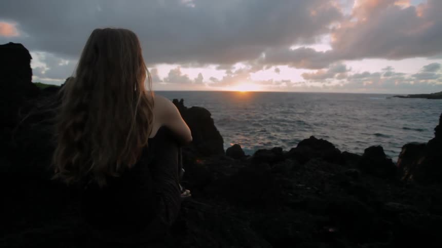 A woman is sitting near the shore at sunset